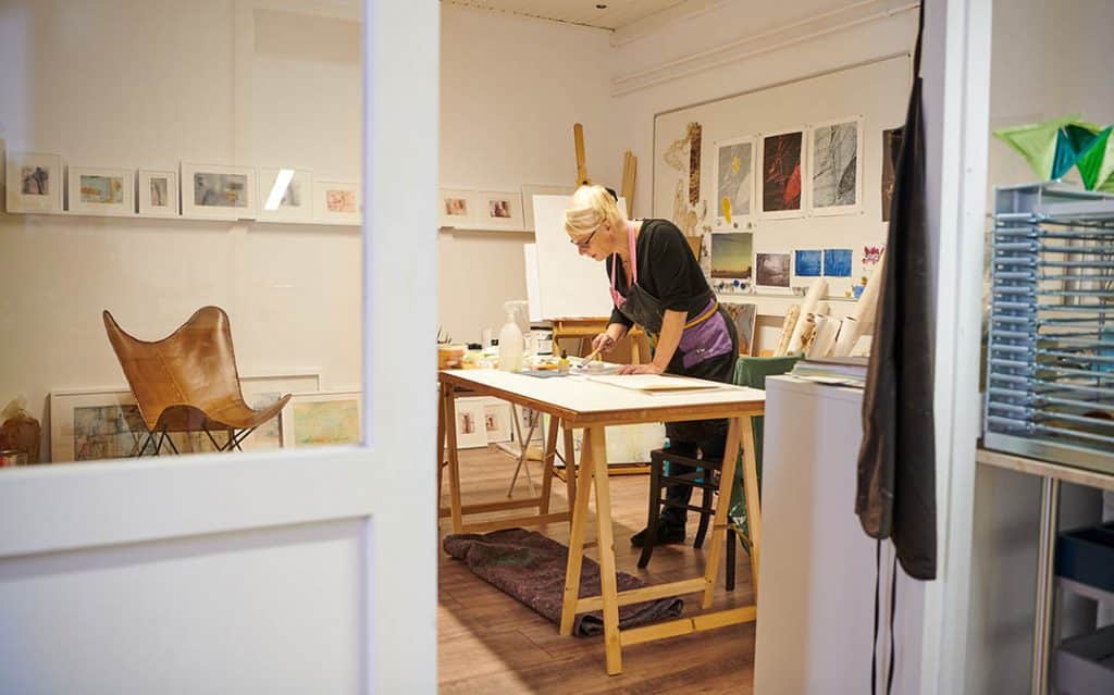 Künstlerin in ihrem Atelier, die hinter einem Arbeitstisch steht und malt. Im Hintergrund sind Kunstwerke zu sehen