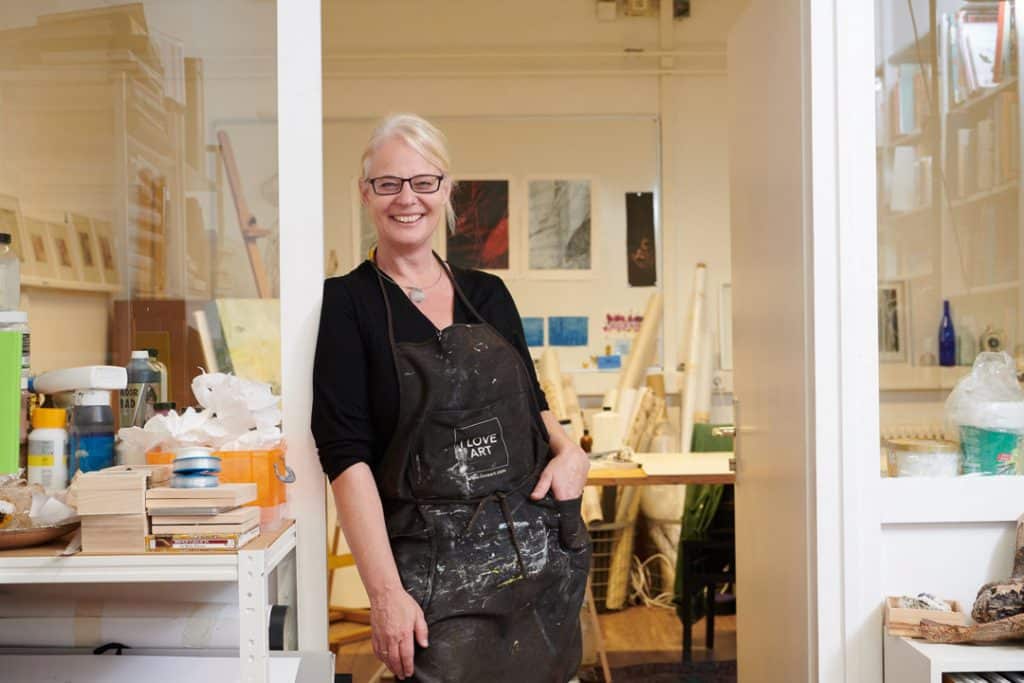 Blonde Frau mit Brille. Künstlerin die in der Tür zu ihrem Atelier steht und in die Kamera lächelt.